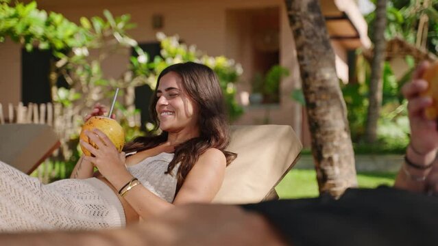 Young happy multiethnic couple relaxing on sunbeds at tropical resort clink and drink coconut water. Cheerful biracial tourists on summer vacation having fresh king coconuts with exotic background