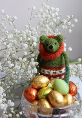 Crochet green toy, chocolate eggs and white flowers on a table. Handmade gift for Easter. Colorful still life photo with oil wrapped sweets. Easter celebration. 
