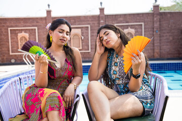 Two Exhausted young Indian women using a paper hand waver fan sitting outdoor in sunny day suffering from hot summer weather or high temperature, Asian Girls get uncomfortable due to heat wave.
