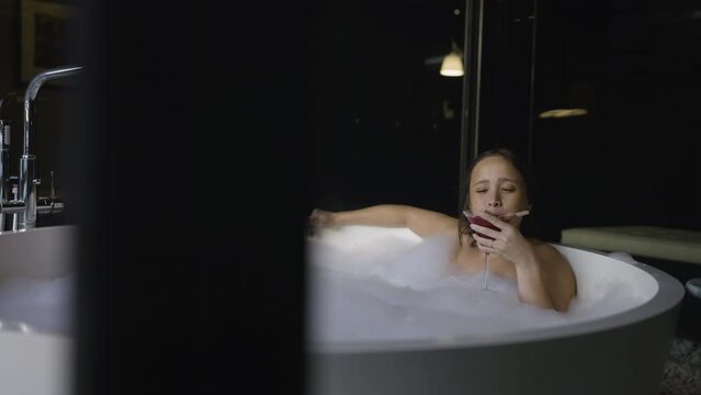 Slow motion shot of an attractive female relaxing in a soapy bath drinking a cocktail