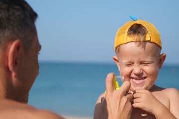 A father applies protective cream to his son's face at the beach. A man's hand applies sunscreen...