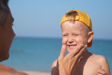A father applies protective cream to his son's face at the beach. A man's hand applies sunscreen...