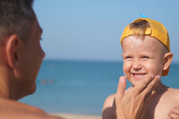 A father applies protective cream to his son's face at the beach. A man's hand applies sunscreen lotion on a child's face. Cute little boy with sunscreen by the sea. Copy space.