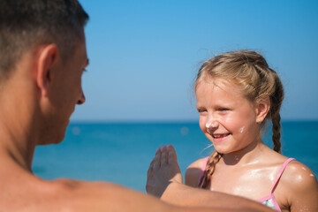 Father applying protective cream to her daughter's face at the beach. Man hand holding sunscreen lotion on baby face. Cute little girl with a sun block by the sea . Copy space.