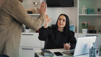 A female executive is sitting with a laptop in her office when her subordinate is brought to her to talk about his success. A woman praises her subordinate and gives him a high five