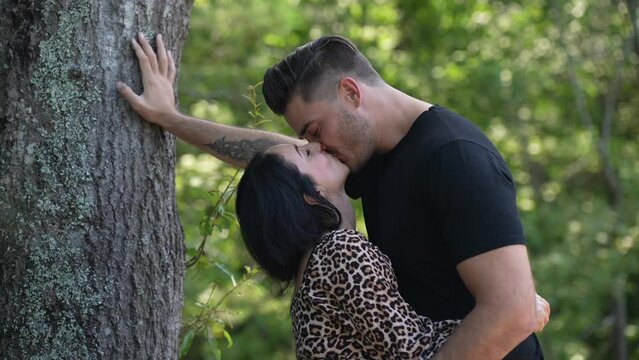 Cute young couple in stylish casual outfits passionately kissing each other outside in nature against a tree and then embracing with a hug, being happy, romantic atmosphere, love story, slow motion