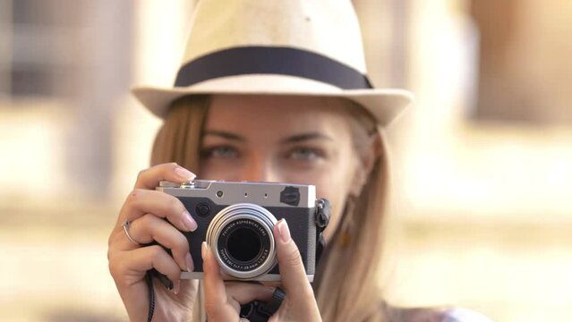Happy young blonde woman with blue eyes and long hair, taking pictures looking at camera with a vintage camera at Colosseum, Rome.
