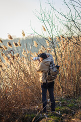 Photographer with camera hiding behind reeds at lake taking pictures of wildlife. Outdoors leisure activity
