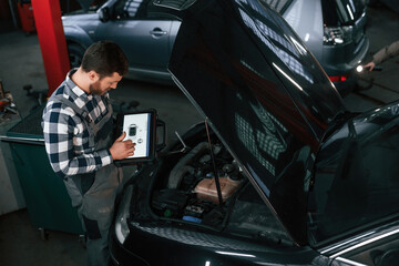 Standing near the car with hood opened. Holding tablet. Man in uniform is working in the auto salon