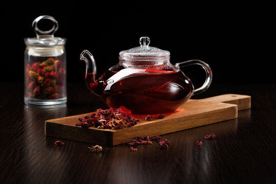 Glass teapot with red herbal tea on a black background. Side view selective focus. Dry rose buds in the background. On the front - dry tea leaves of hibiscus.