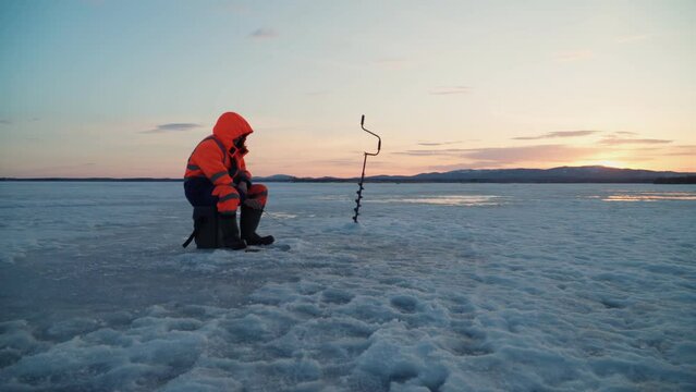 A fisherman catches fish on the ice of a frozen lake at sunset. Winter fishing and ice fishing.