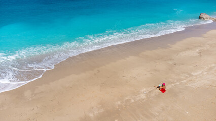 Fototapeta na wymiar Aerial view of woman in red dress sitting on the sandy beach with a baby, enjoying soft turquoise ocean wave. Tropical sea in summer season on Megali Petra beach on Lefkada island.