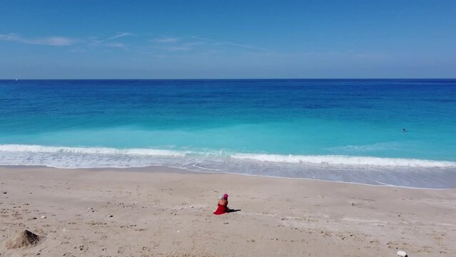Aerial view of woman in red dress sitting on the sandy beach with a baby, enjoying soft turquoise ocean wave. Tropical sea in summer season on Megali Petra beach on Lefkada island.