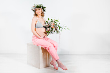 Pregnant woman in wreath with bouquet of flowers. Girl in gray top, pink pants. Hands on naked belly. Photoshoot happy pregnancy,maternity,preparation. Baby expectation. Beautiful tender young mother