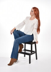 full length portrait of beautiful woman model with long red hair, wearing casual outfit white blouse  top and denim jeans, isolated on white studio background. Relaxed seated pose, sitting on chair wi