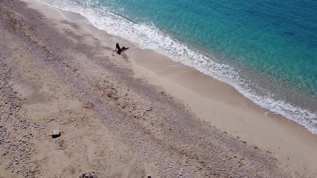 Aerial view of beautiful happy woman in swimsuit sitting on sand in the shallow sea water, enjoying beach and turquoise ocean wave. Tropical sea in summer season on Egremni beach on Lefkada island.