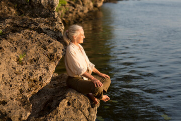 Summer lifestyle portrait senior woman with gray hair relaxes sitting on rocks on the seashore. Enjoying the little things. spends time in nature in summer.  meditation.