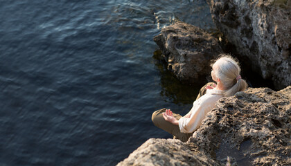Summer lifestyle portrait senior woman with gray hair relaxes sitting on rocks on the seashore. Enjoying the little things. spends time in nature in summer.  meditation.