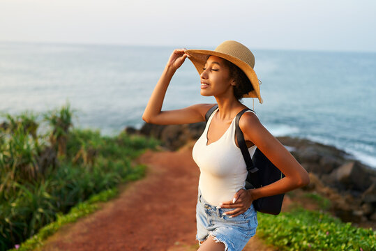Multiethnic woman in straw hat enjoys tropical vacation travel hike. Black female with backpack sightseeing on scenic location. Pretty lady standing on island on sunrise. Girl in the sun for suntan