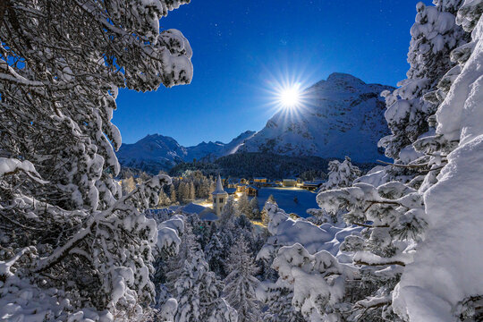 Moon glowing over the snowy bell tower of Chiesa Bianca and woods in winter, Maloja, Bregaglia, Engadine, Canton of Graubunden, Switzerland