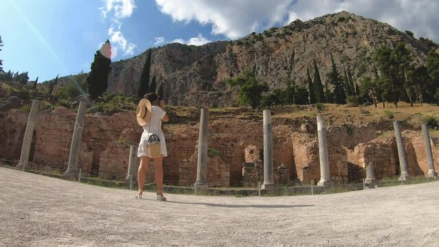 Young woman taking pictures at ruins of temple of Apollo in Delphi, archaeological site by mount Parnassus, Greece, Europe. Vintage camera, large hat, fashion white dress.