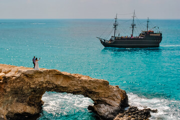A couple of newlyweds, a bride and groom, stand on the rocky Love Bridge in Cyprus, near Ayia Napa. Against the background, a retro-style ship sails through the sea