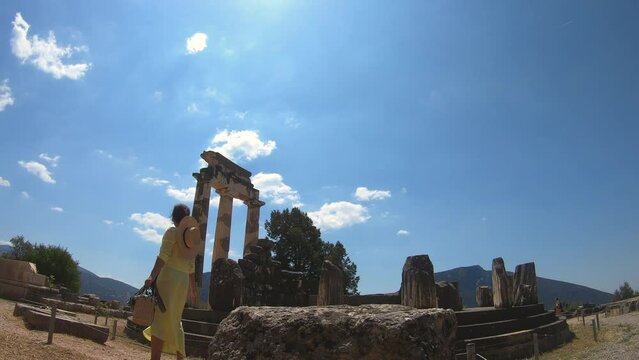 Young woman tourist visiting temple of Athena Pronaia in archaeological site of Delphi, Voiotia, Greece. Fashion elegant yellow dress, large hat.