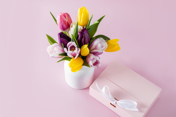 A lovely bouquet of colorful tulips in a white ceramic vase and a gift pink box on a pastel background. festive composition.