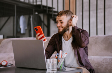 Puzzled caucasian beardy caucasian man in casual sitting on couch at desk with laptop holds phone...