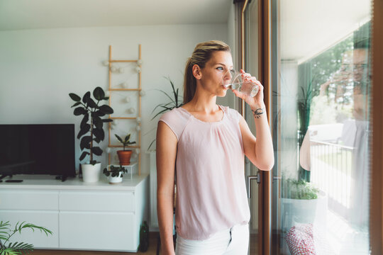 Blonde woman taking her supplements with a glass of water, standing by the window looking out