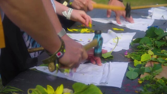 group of adults and children are bashing hitting leaves with hammer placed under a piece of cloth to create artistic piece out of it close up on hands flowers on table family are on music festival