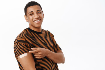 Fototapeta na wymiar Health and vaccination concept. Smiling healthy african american man, 30 years, has band aid on shoulder after covid vaccine shot, white background