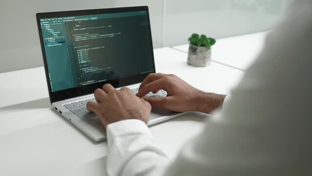 Over-the-shoulder view of qualified male programmer coding on laptop in bright modern IT company developing. Professional IT worker typing on computer working doing tasks.