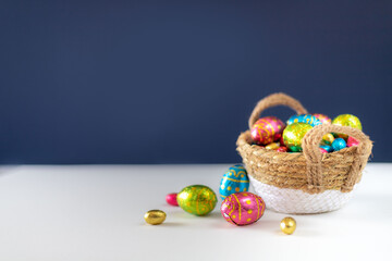 Fototapeta na wymiar Easter eggs in basket with blue background on white table. Studio lighting colorful easter decorated eggs for egg hunt with wicker basket and flowers. Banner for web and composition. Wishes card