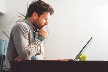 Side view waist up bearded caucasian man sitting by the table looking at his laptop, online...