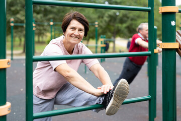 Aged woman doing stretching on sports bars in open-air sports area