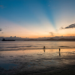 Sunset at the beach. Silhouette of people near the sea and a cargo ship passing by on the horizon. Long exposure photography. Santos, Brazil.