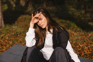 Sad woman sitting on yoga mat in the autumn park and look sad, she hold her head hand, drama queen, depression or bad mood, problems. Woman wear white long sleeves and black waistcoat.