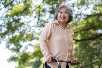 Happy old elderly Asian woman uses a walker for osteoarthritis rehabilitation physiotherapy in park.  Concept of happy retirement With care from a caregiver and Savings and senior health insurance