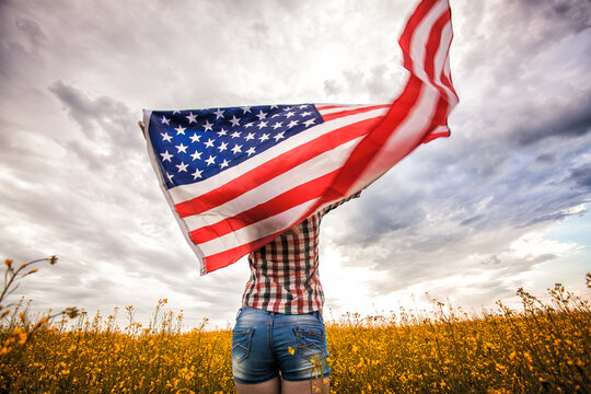 Beautiful young girl holding an American flag on the wind in a rapeseed field. Summer landscape against the blue sky.
