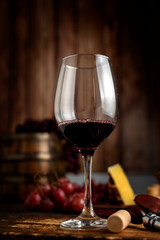 red wine glass with bottle serving wine on still life table with wooden barrel grapes and cheese on wooden boards