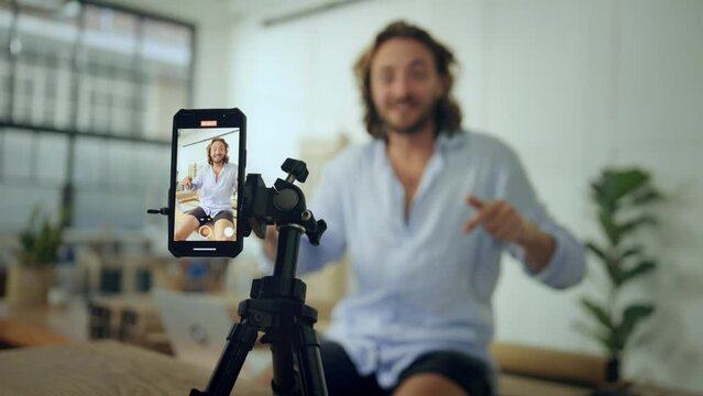Young caucasian man in shirt gesturing while sitting and talking on video by mobile phone in office