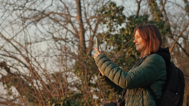 A young adult caucasian woman backpacker taking a picture of a landscape with her phone at the sunset