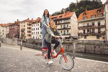 Fototapeta premium Couple of best friends riding a bike on the city streets, one of the women standing on the trunk of the bike 