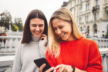 Waist up portrait two smiling young women in red and beige sweater walking around the city looking down at their phone 