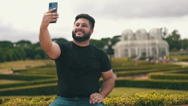 Young man picking up his cell phone and taking a selfie at the park outdoors while seated on a long wood bench	located at Botanical Garden, Curitiba, Brazil