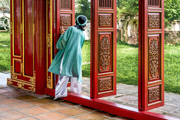 Naklejka premium Vietnam, Lady in traditional dress in the Red wooden doorway in the To Mieu Temple a building in the Forbidden City of the Imperial City in the Citadel of Hue.