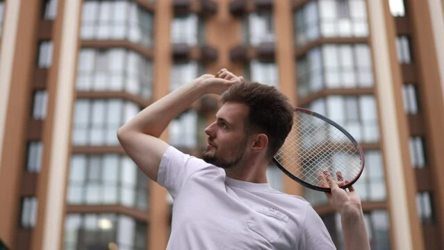 Young confident sportsman with tennis racket behind head standing at background of urban residential building outdoors. Portrait of fit handsome Caucasian player with equipment looking around