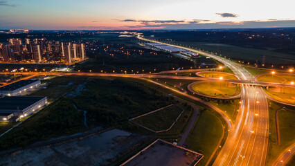 Aerial view of the bigger transportation interchange with many cars next to forest and the historical and at same time modern city of St. Petersburg at light summer night, fog