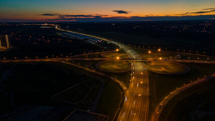 Aerial view of the bigger transportation interchange with many cars next to forest and the historical and at same time modern city of St. Petersburg at light summer night